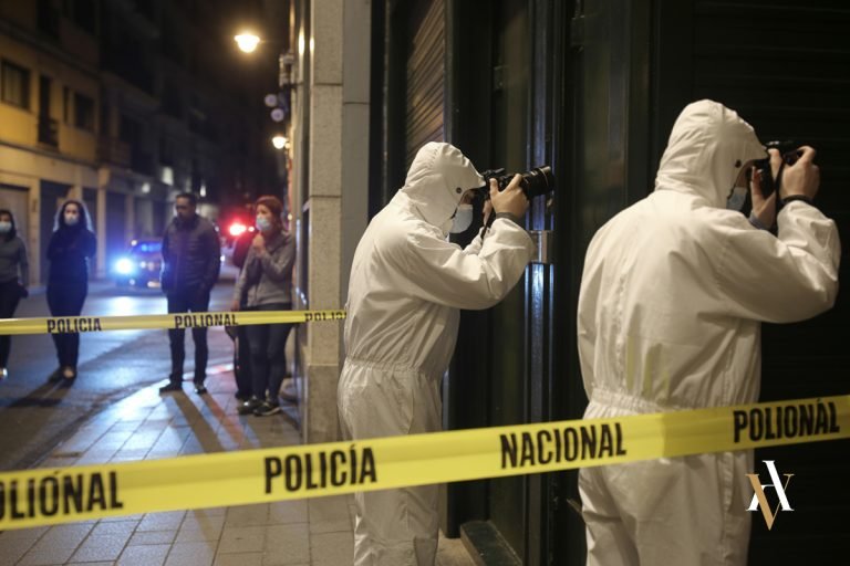 Agentes de la Policía Nacional y forenses fotografiando una escena del crimen durante una investigación policial penal en una calle urbana de España, con el área acordonada y varios testigos observando a lo lejos.