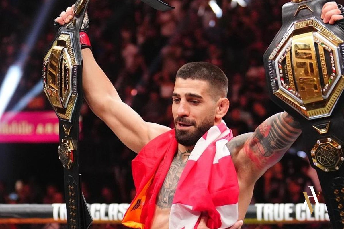 Ilia Topuria celebrando una victoria en la UFC con dos cinturones de campeón, sosteniéndolos en alto dentro del octágono
